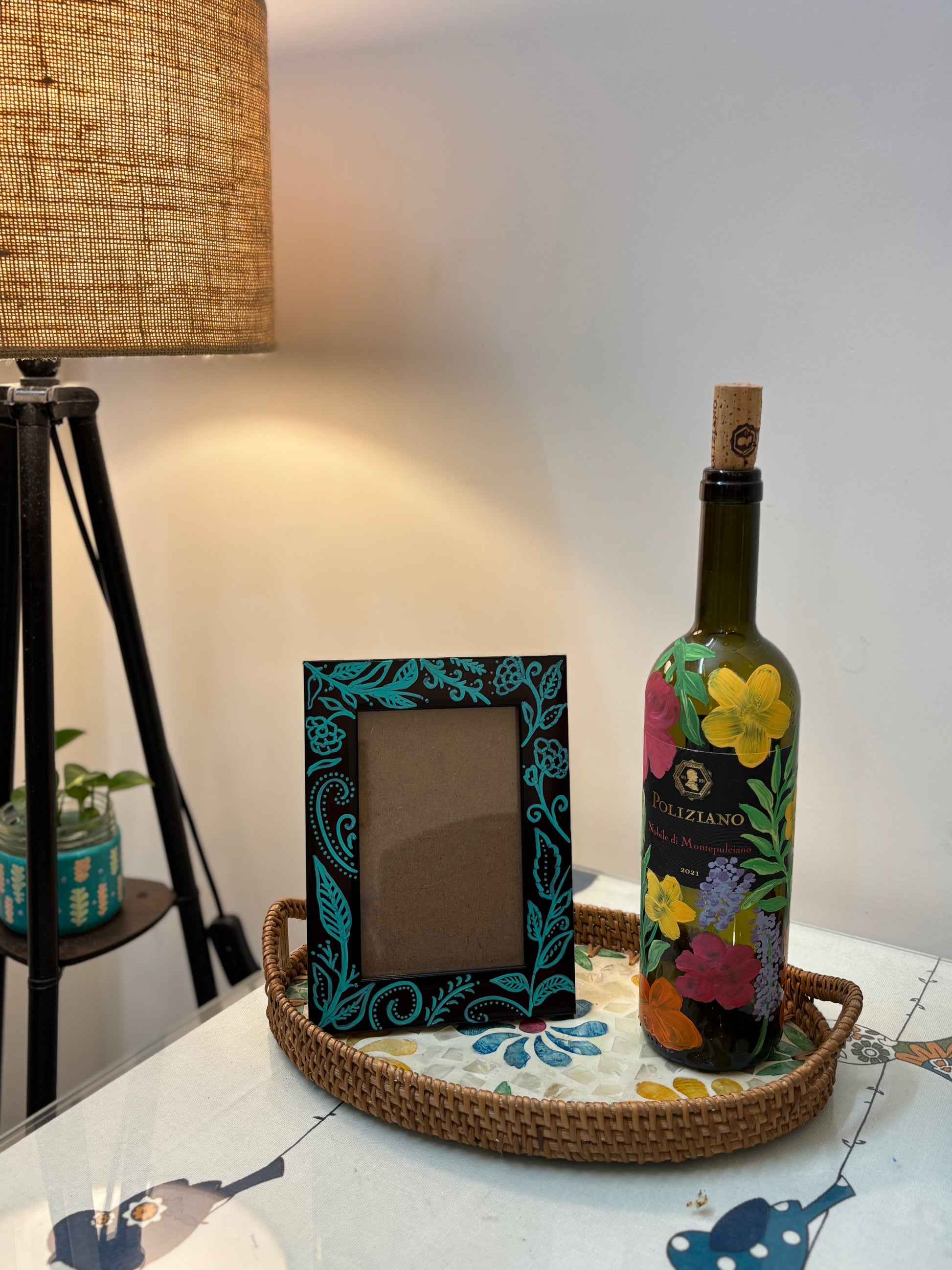 Decorative bottle with floral design on a tray next to a framed picture on a table. kalavrit hand painted frame