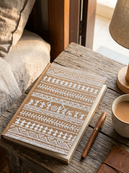 A hardcover notebook with a beige cover and white geometric tribal-style patterns, resting on a rustic wooden table next to a cup of coffee, a pen, and a lamp.